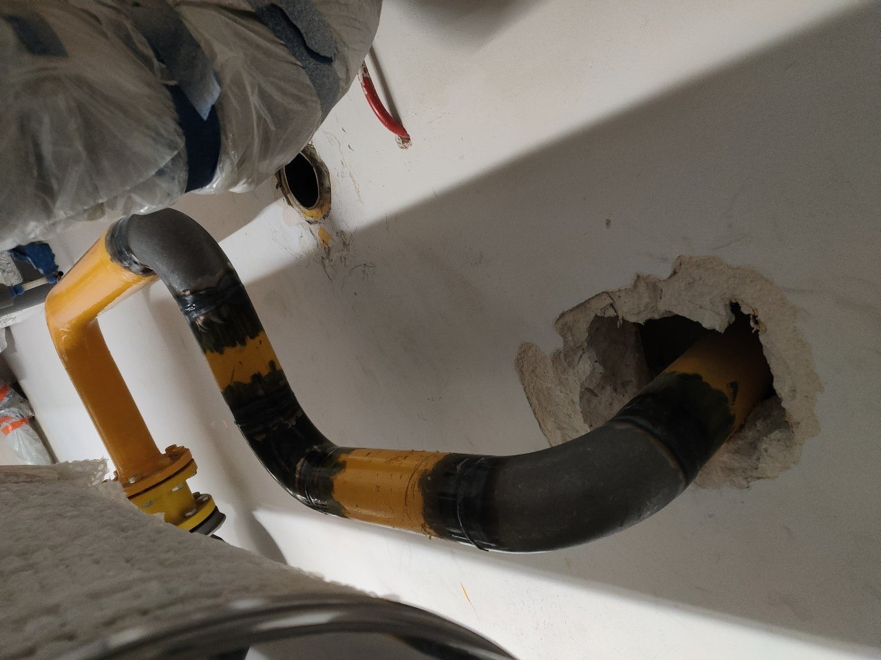 Żółto-czarna rura gazowa, częściowo zaizolowana, przechodzi przez otwór w ścianie. Widoczne łączenia i elementy instalacji. Ujęcie z bliska, z dołu.