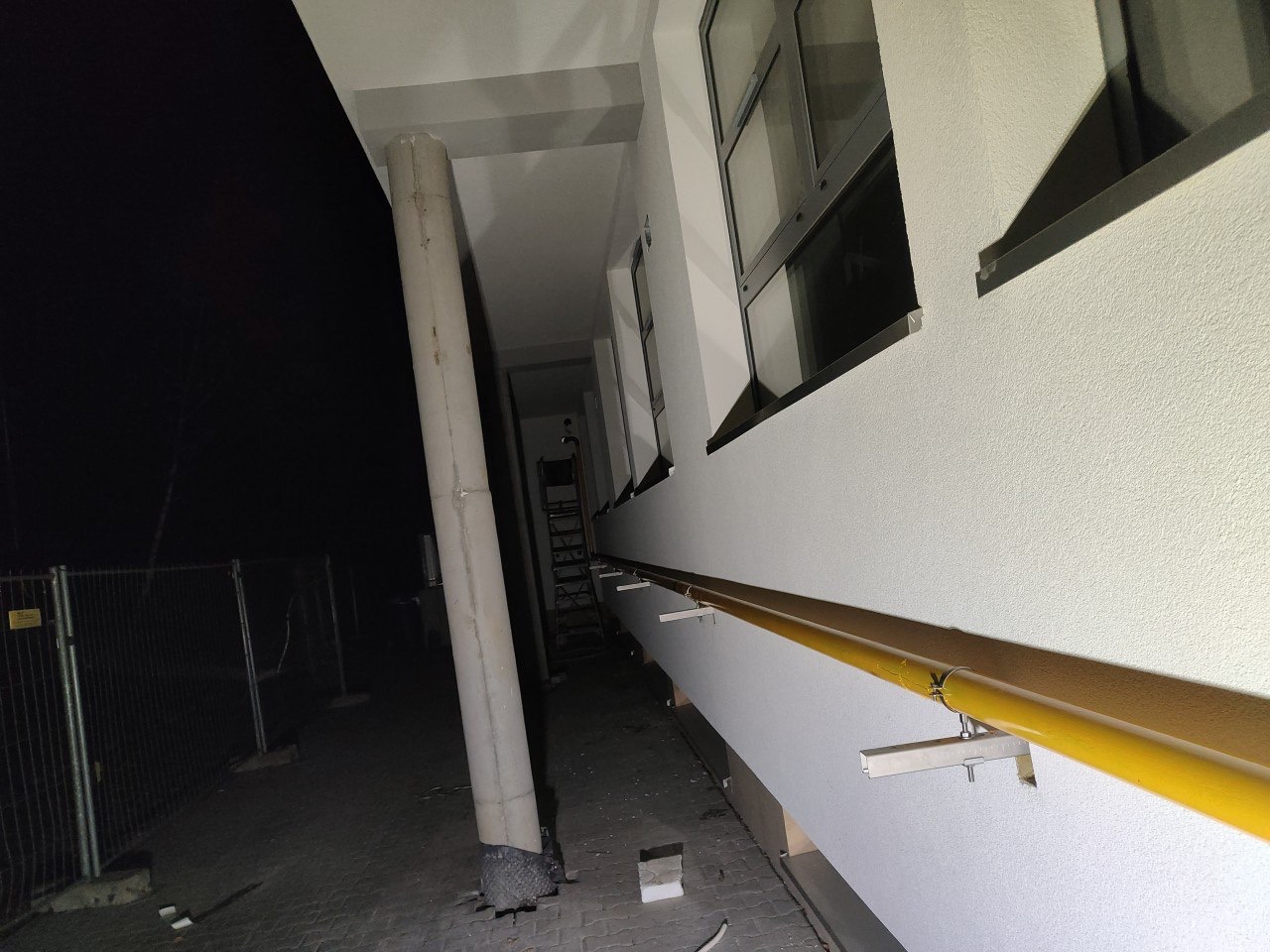Zewnętrzny korytarz budynku w nocnej scenerii. Widoczny betonowy filar, żółta poręcz i okna. W tle siatka ogrodzeniowa i drabina.