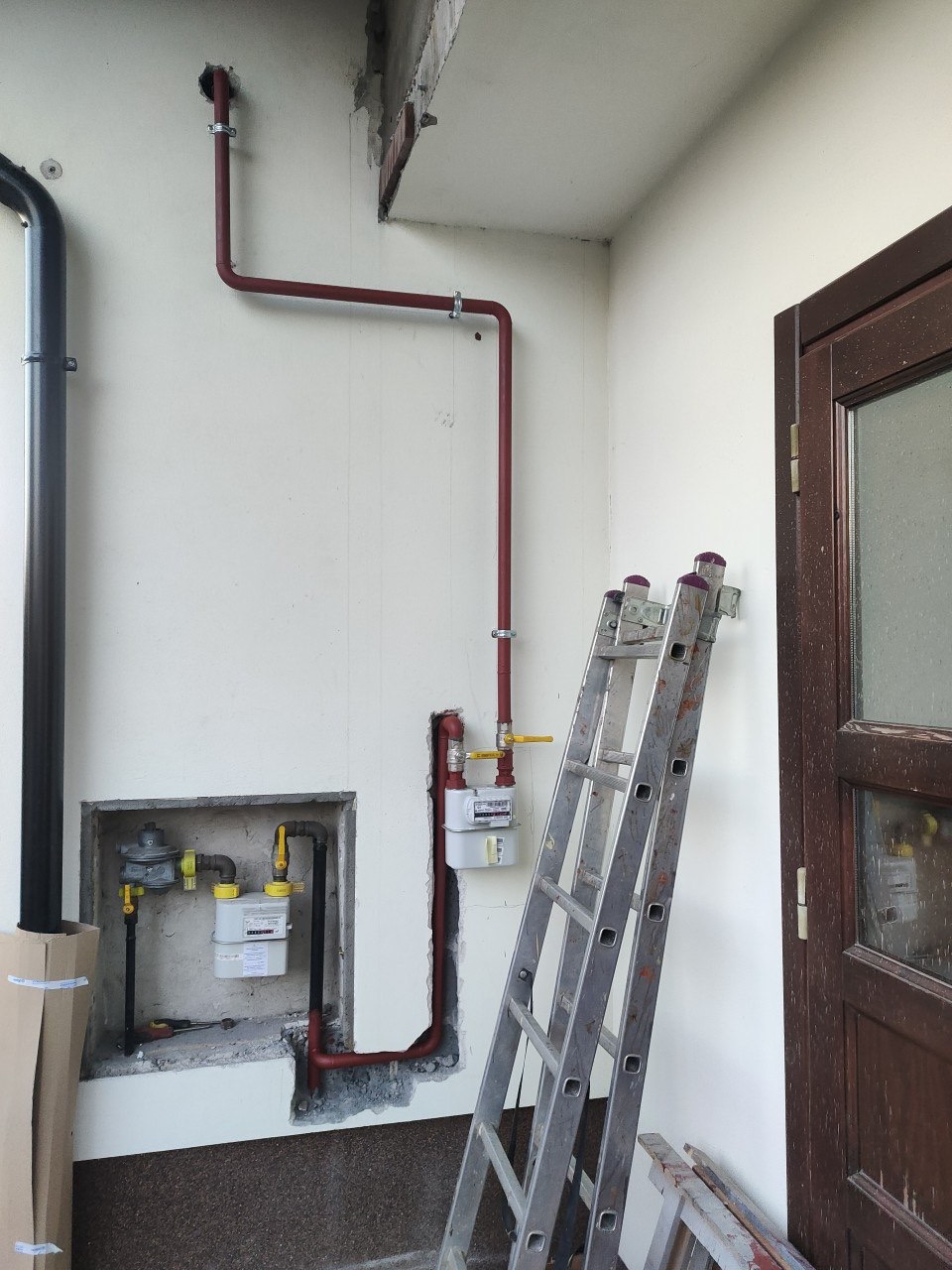 Czerwona instalacja gazowa z licznikiem, zamontowana na białej ścianie w rogu pomieszczenia. Obok drabina aluminiowa i brązowe drzwi. Widoczne żółte zawory.