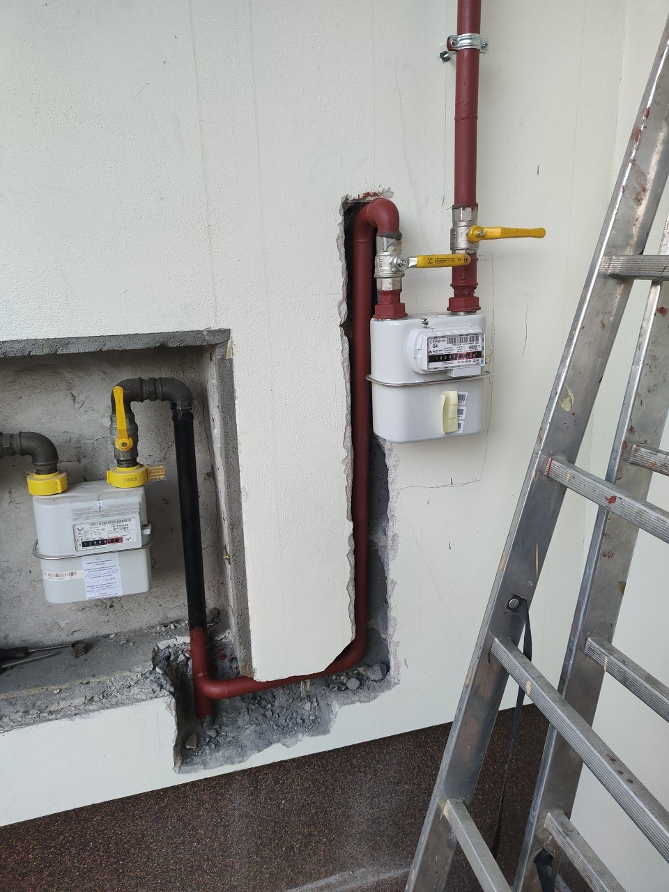 Dwa gazomierze z rurami, jeden z czerwoną rurą pionową, drugi z czarną, oba z żółtymi zaworami, zamontowane na białej ścianie. Widoczna drabina.