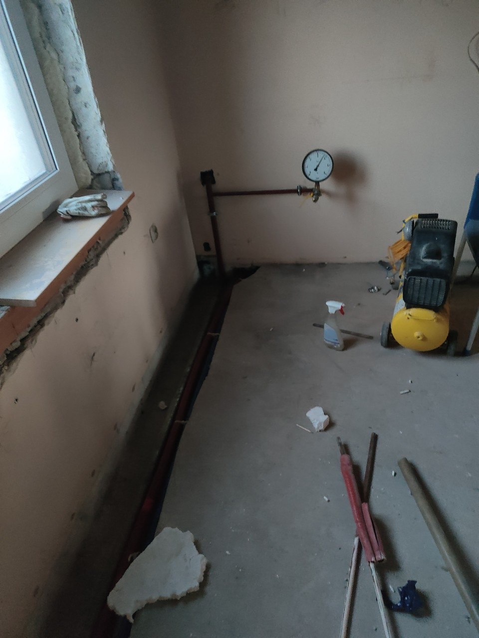 Instalacja gazowa w trakcie prac: rury w bruzdach, manometr na ścianie, kompresor obok. Widoczne narzędzia i resztki materiałów budowlanych na surowej posadzce.