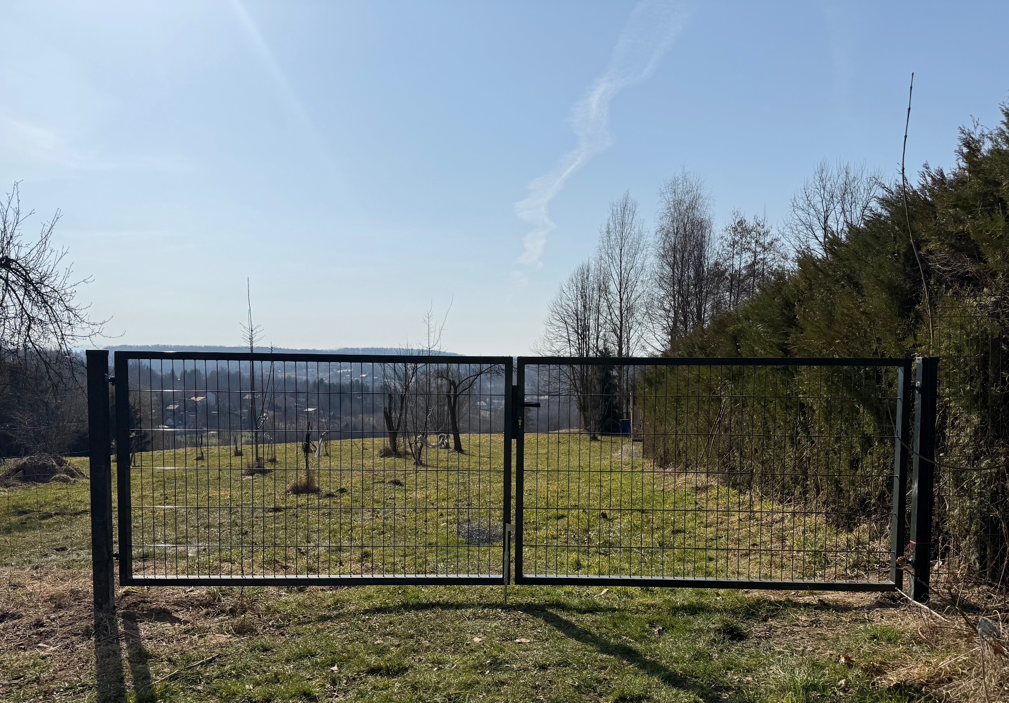 Nowoczesna, dwuskrzydłowa brama panelowa w kolorze grafitowym, z widokiem na pagórkowaty krajobraz i błękitne niebo z delikatną smugą kondensacyjną.