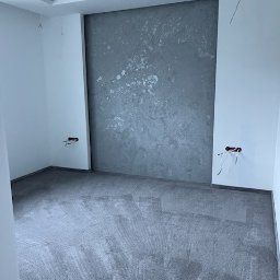 DECOR SYSTEM DESIGN SPÓŁKA Z OGRANICZONĄ ODPOWIEDZIALNOŚCIĄ - Pokój po szpachlowaniu: szara ściana z efektem dekoracyjnym, przygotowana podłoga, wystające kable elektryczne. Widok z niskiego kąta.