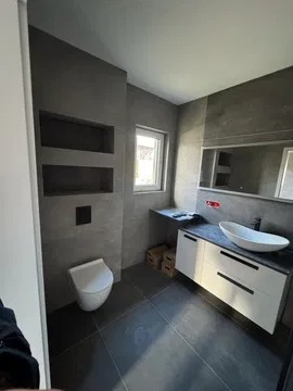 Nowoczesna łazienka w szarościach: podwieszana toaleta, umywalka nablatowa, szafka z białymi frontami, wnęki w ścianie. Minimalistyczny design, ciemna podłoga.