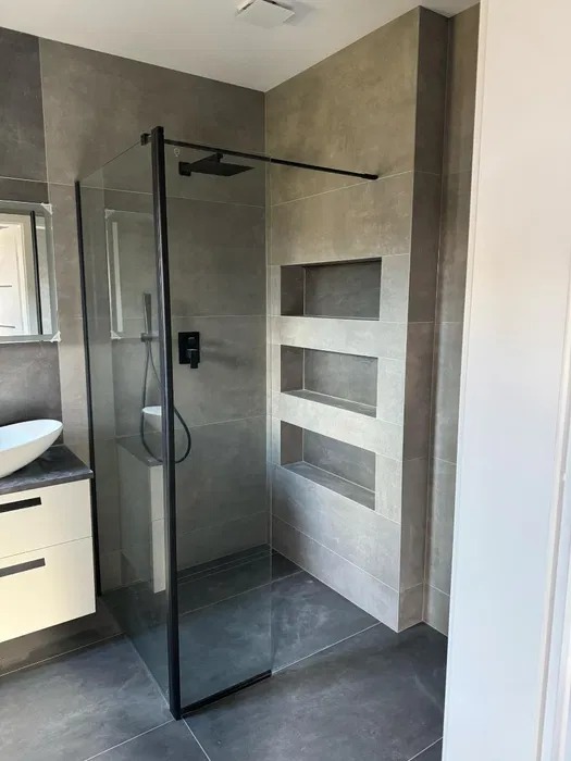 Nowoczesna łazienka z kabiną typu walk-in, szare płytki i wnęki ścienne. Minimalistyczny design z czarną armaturą i umywalką nablatową.