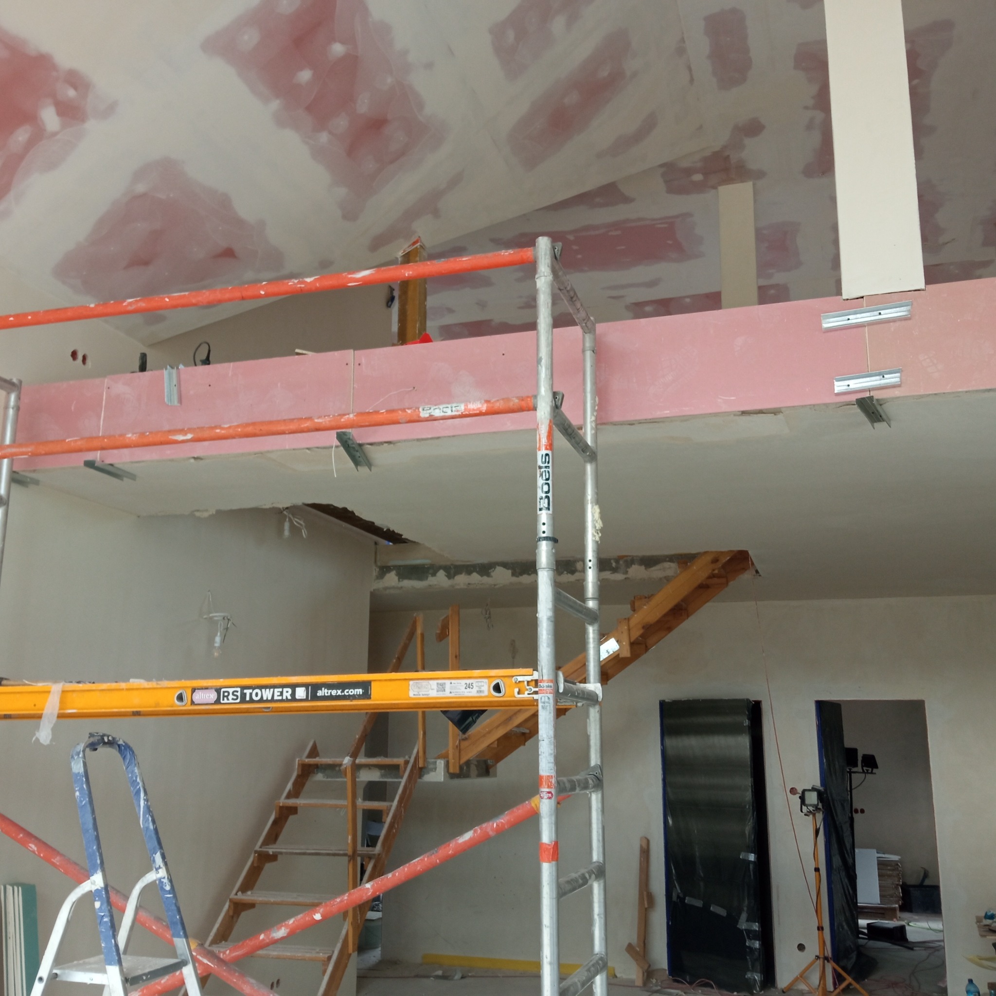 Sufit z płyt kartonowo-gipsowych w trakcie szpachlowania, widoczne rusztowania i przygotowane otwory drzwiowe. Prace wykończeniowe w surowym wnętrzu.