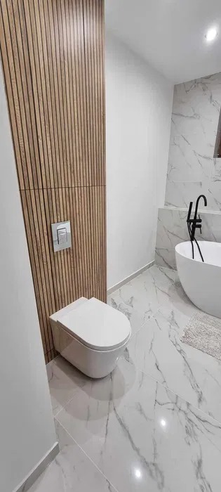 Nowoczesna łazienka z drewnianą ścianą za toaletą, marmurową podłogą i wolnostojącą wanną z czarną armaturą. Minimalistyczny design, jasne wnętrze.