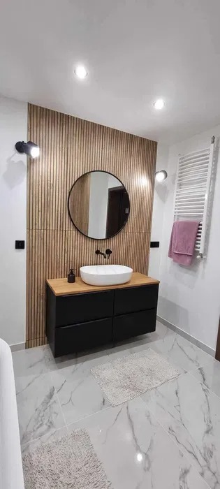 Nowoczesna łazienka w Warszawie: drewniane lamele na ścianie za okrągłym lustrem, biała umywalka nablatowa na czarnej szafce, marmurowe płytki podłogowe.