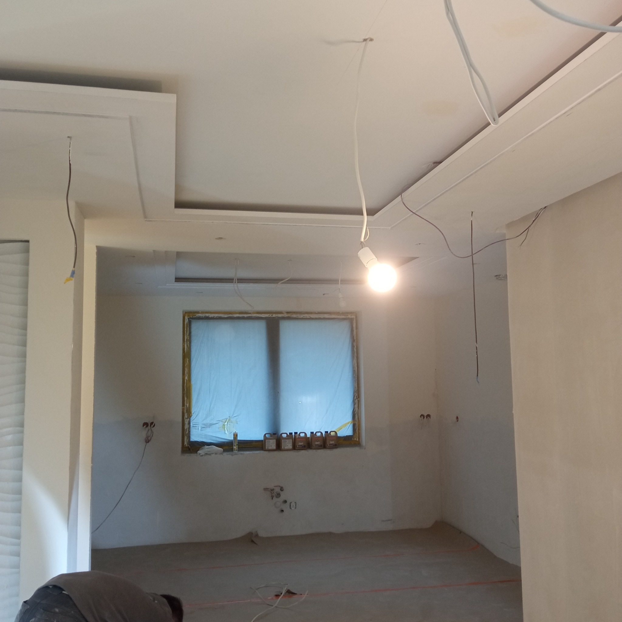 Wnętrze w trakcie wykończenia: podwieszany sufit z oświetleniem, okno zabezpieczone folią, widoczne przewody elektryczne. Etap prac wykończeniowych.