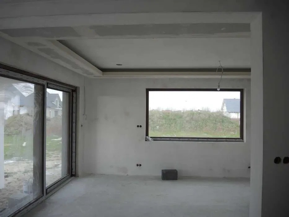 Surowe wnętrze z oknem i przesuwnymi drzwiami balkonowymi. Ściany przygotowane do malowania, widoczne ślady szpachlowania i płyty karton-gips na suficie.