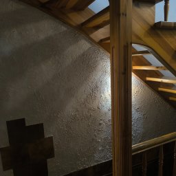 Usługi Budowlane Karol Krawczyk - Ściana z dekoracyjną fakturą i drewniane schody z balustradą. Widoczne detale wykończenia wnętrza, oświetlenie podkreśla strukturę ściany i naturalne drewno.
