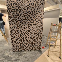 Usługi Budowlane Karol Krawczyk - Montaż tapety w panterkę na ścianie. Widoczna drabina, narzędzia i fragment osoby montującej. Podłoga wyłożona płytkami imitującymi beton.