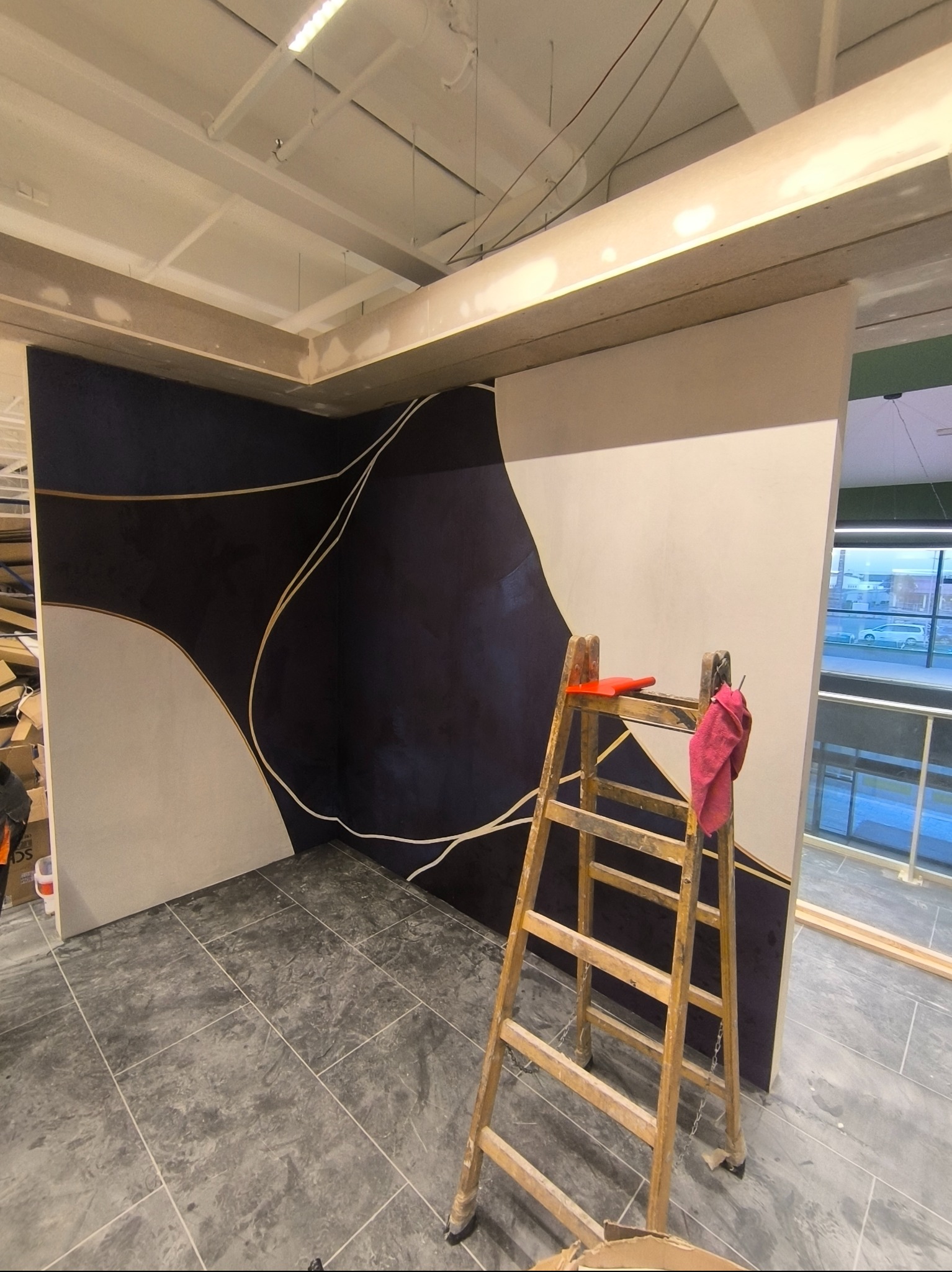 Wykończona ściana z abstrakcyjnym wzorem w odcieniach granatu i beżu, z drewnianą drabiną i szmatką w pomieszczeniu z szarą podłogą. Widoczne niedokończone elementy sufitu.