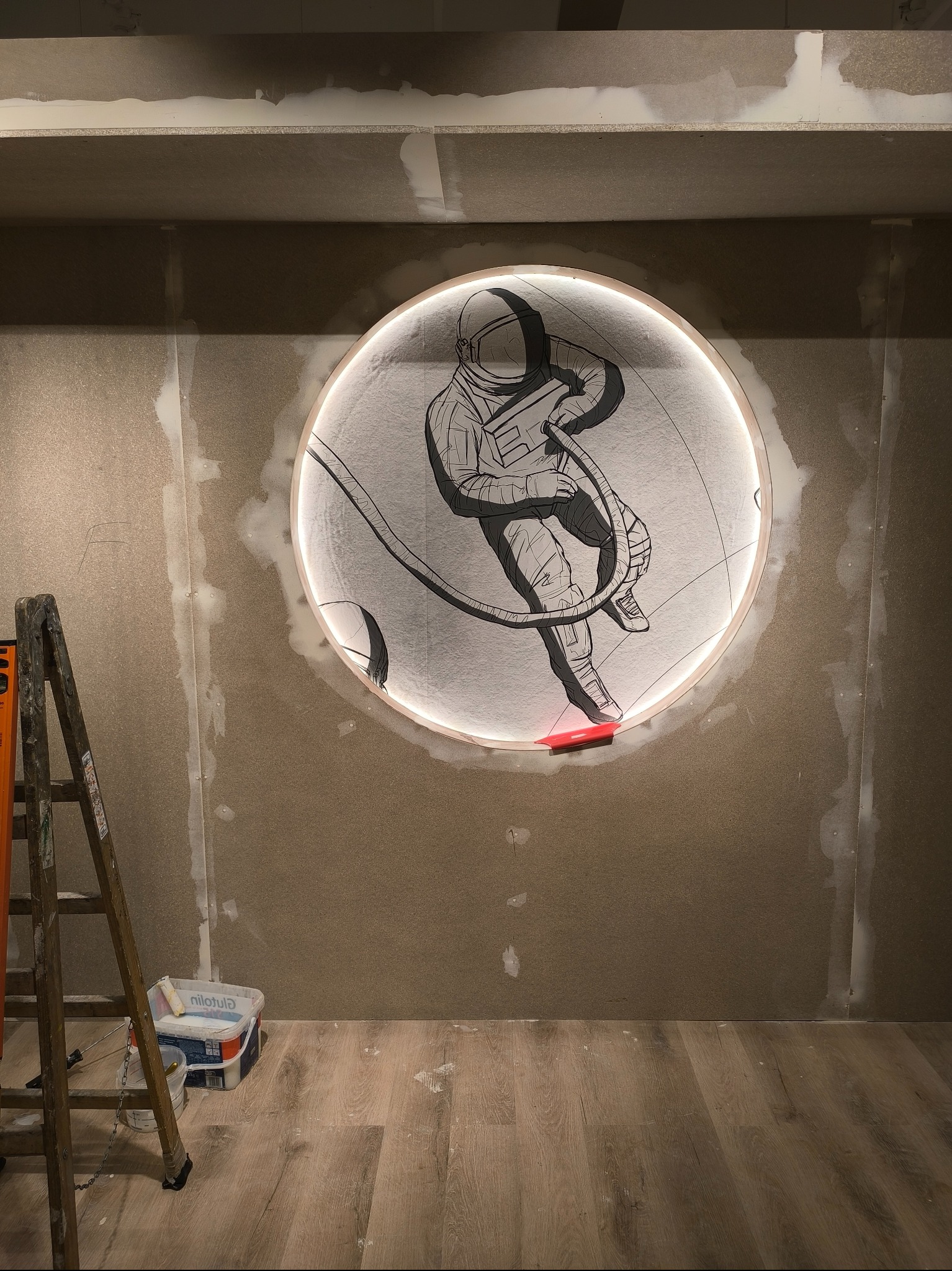Montaż okrągłej dekoracji ściennej z motywem astronauty w Sławie. Widoczne ślady szpachlowania i drabina, co wskazuje na etap prac wykończeniowych.