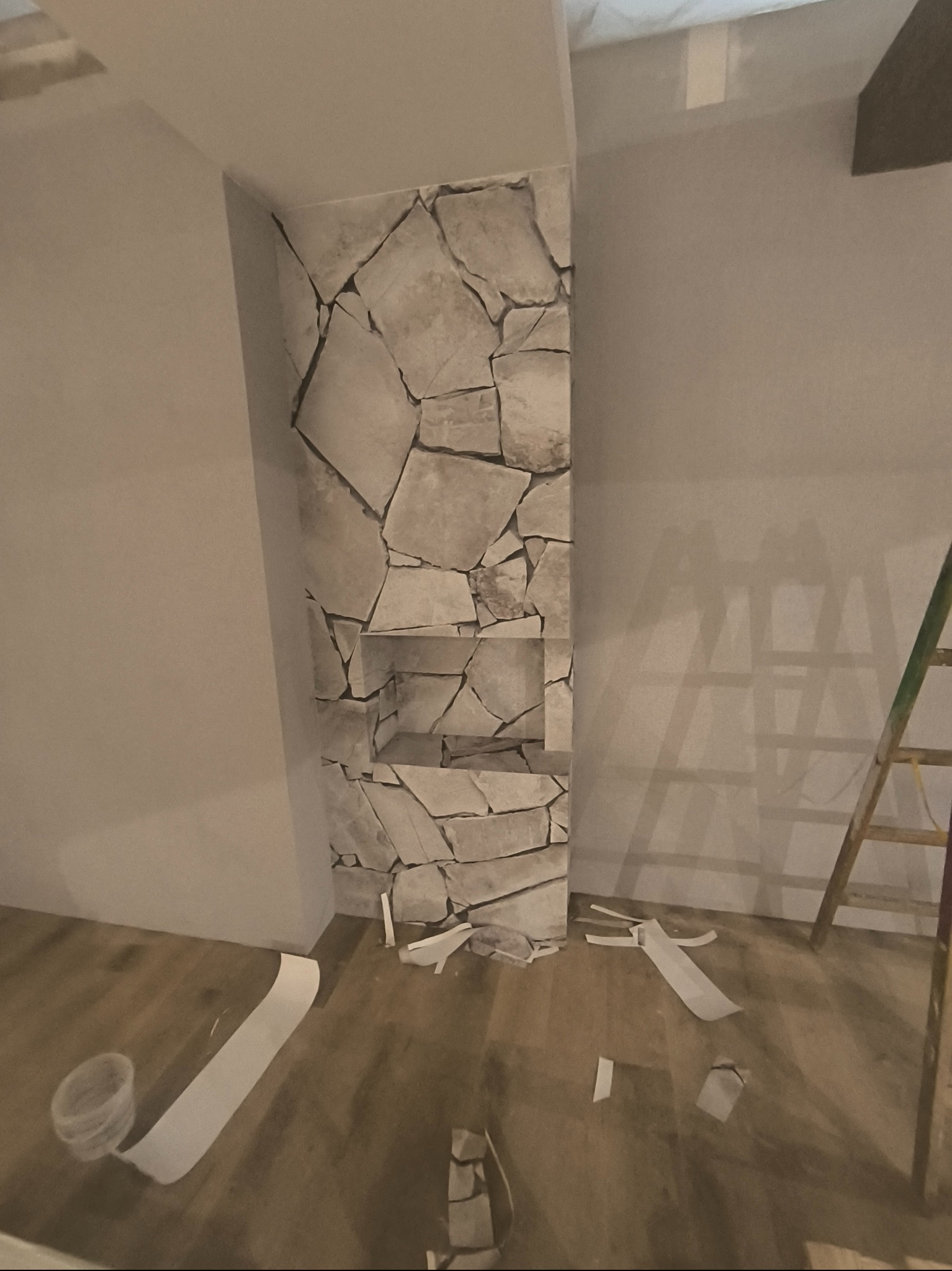Narożnik ściany z wnękami wykończony tapetą imitującą kamień, fragment drabiny malarskiej, resztki tapety na podłodze. Ujęcie z bliska, w trakcie prac.