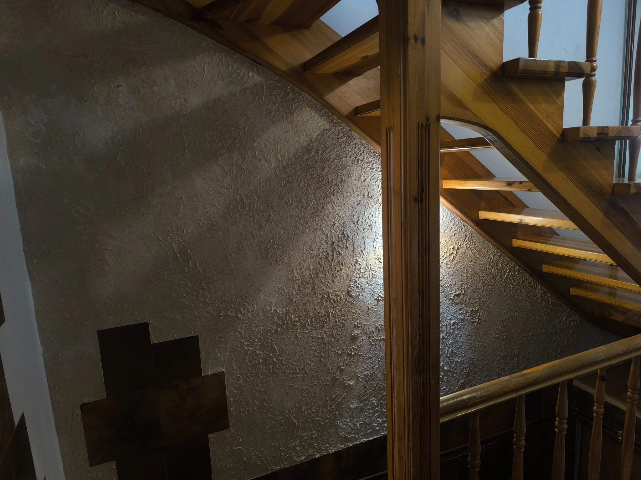 Ściana z dekoracyjną fakturą i drewniane schody z balustradą. Widoczne detale wykończenia wnętrza, oświetlenie podkreśla strukturę ściany i naturalne drewno.