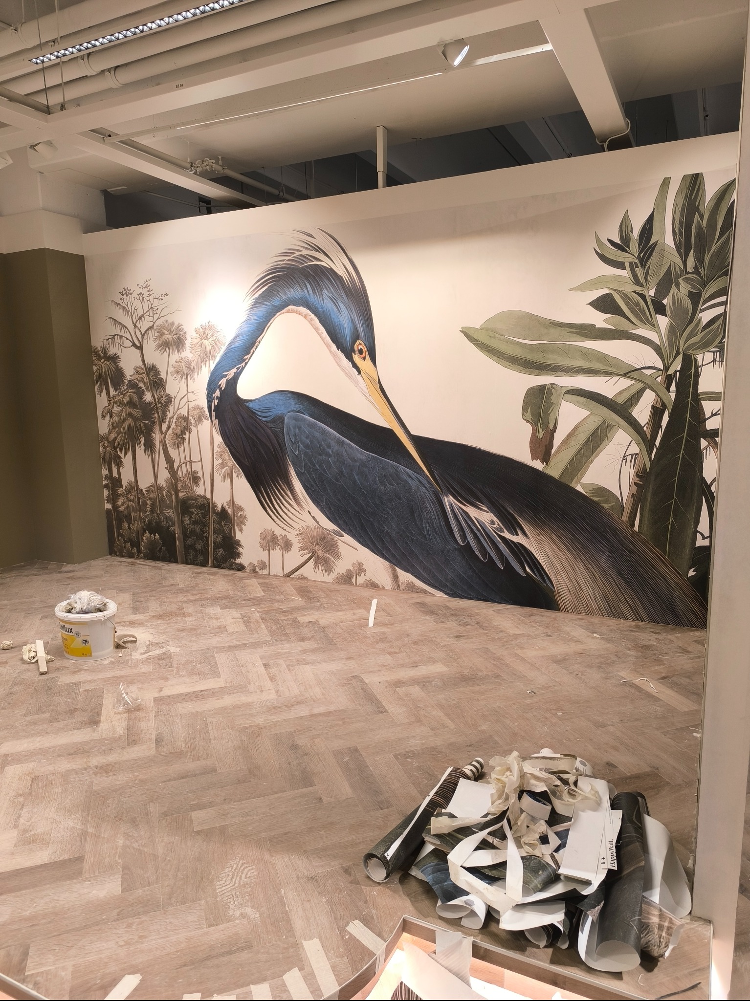 Pokój z tapetą z motywem czapli i tropikalnych roślin. Na podłodze resztki tapety i wiadro z klejem. Widoczny fragment parkietu ułożonego w jodełkę.