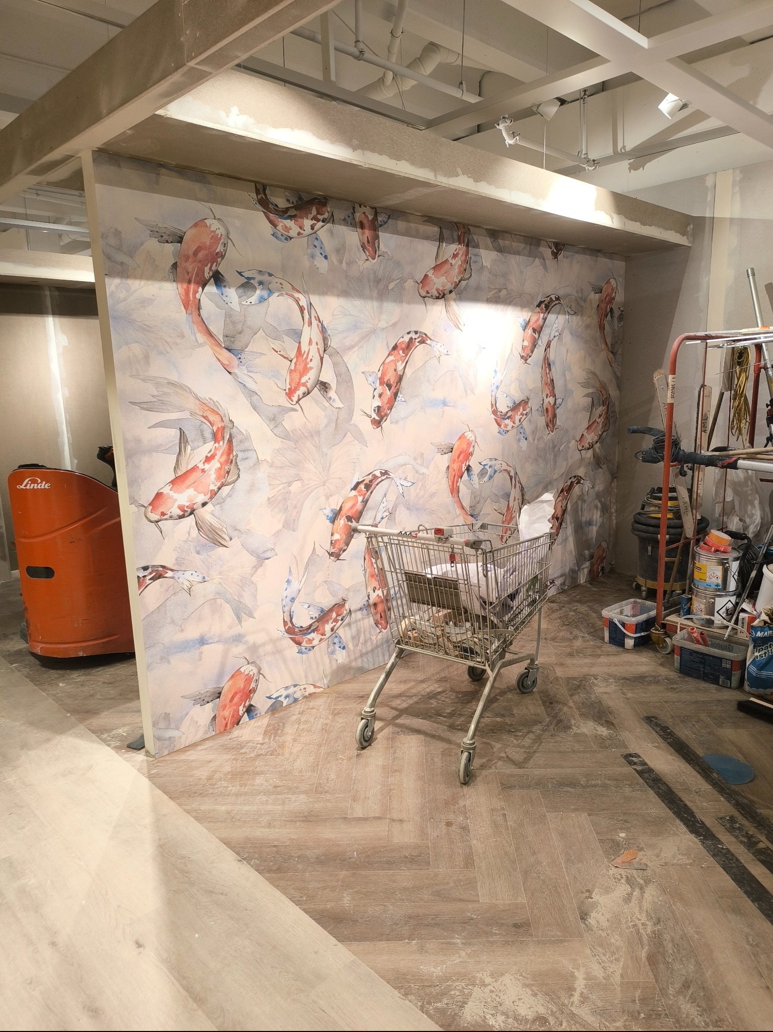 Ściana z tapetą w motywy koi, w trakcie prac wykończeniowych. W tle wózek z narzędziami i podnośnik. Podłoga wyłożona panelami w jodełkę.