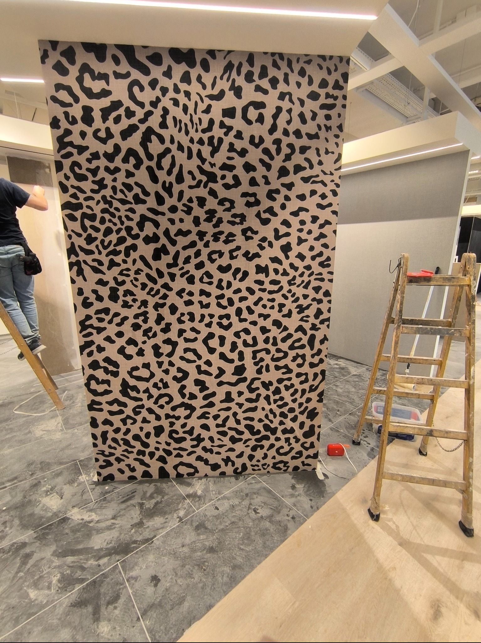 Montaż tapety w panterkę na ścianie. Widoczna drabina, narzędzia i fragment osoby montującej. Podłoga wyłożona płytkami imitującymi beton.