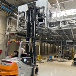 IZI inż. Michał Syncerz - Wózek widłowy STILL podnosi moduł elektryczny w hali produkcyjnej w Jędrzejowie. Widoczne instalacje pod sufitem i oświetlenie. Operator w kabinie.