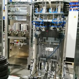 IZI inż. Michał Syncerz - Otwarta szafa sterownicza z widocznymi elementami elektrycznymi, pneumatycznymi i hydraulicznymi. Skomplikowany układ przewodów i komponentów. Widoczne ostrzeżenie o napięciu.