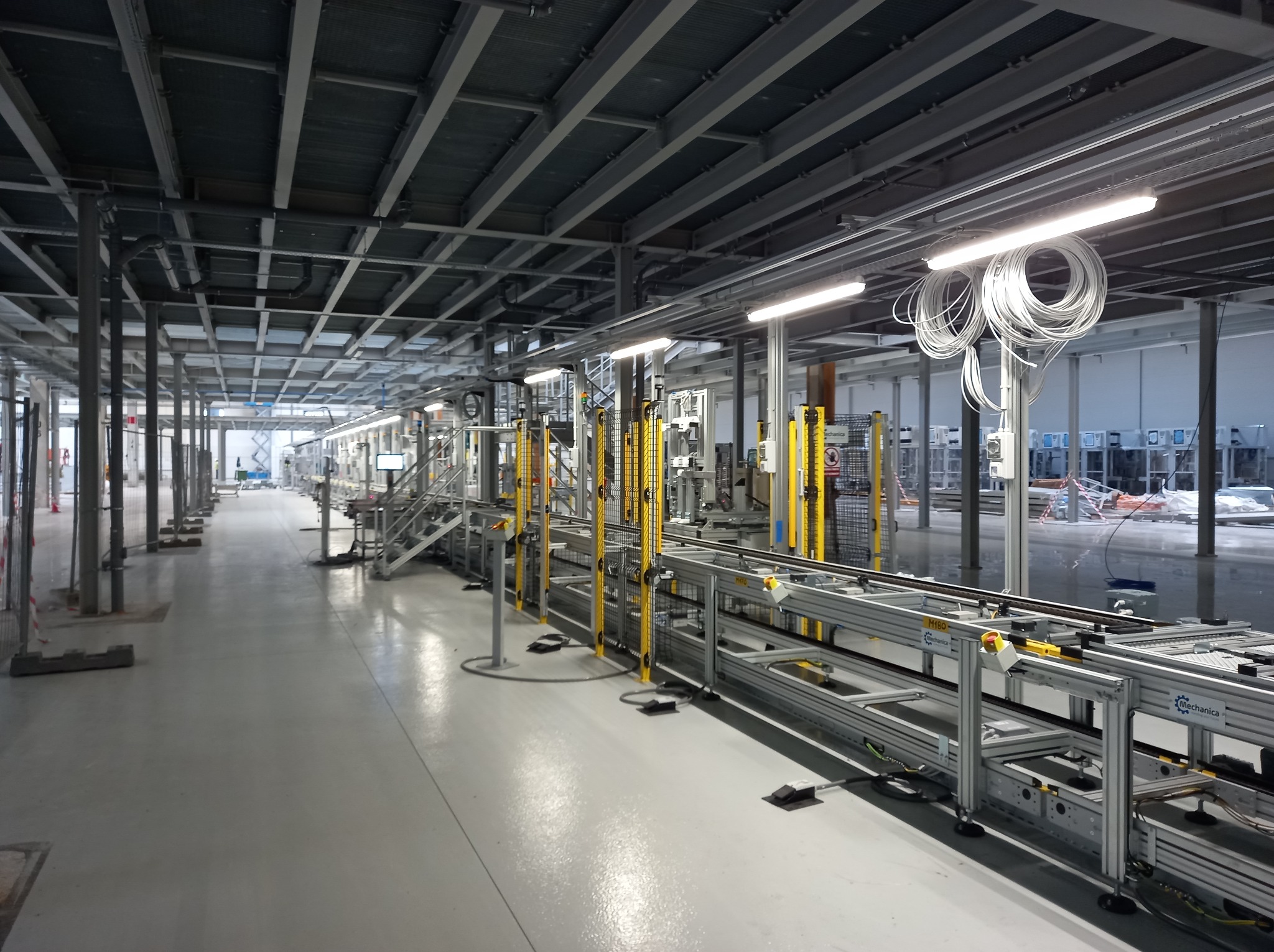 Nowoczesna hala produkcyjna z systemem transportu wewnętrznego. Widoczne metalowe konstrukcje, oświetlenie LED i instalacje pod sufitem. Perspektywa w głąb hali.