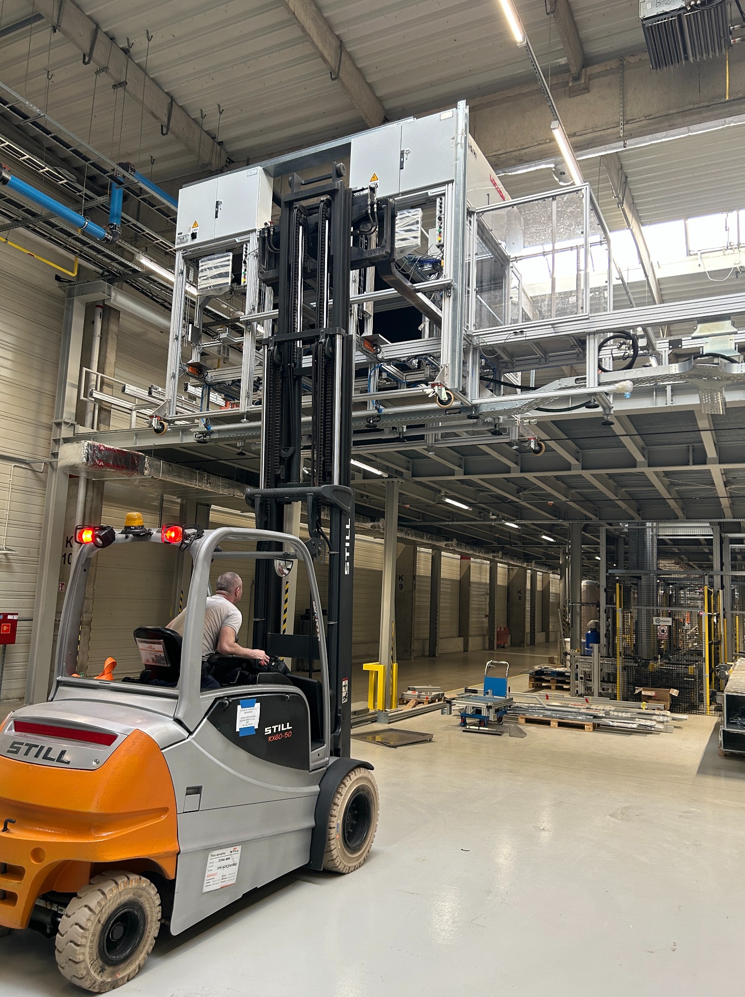 Wózek widłowy STILL podnosi moduł elektryczny w hali produkcyjnej w Jędrzejowie. Widoczne instalacje pod sufitem i oświetlenie. Operator w kabinie.