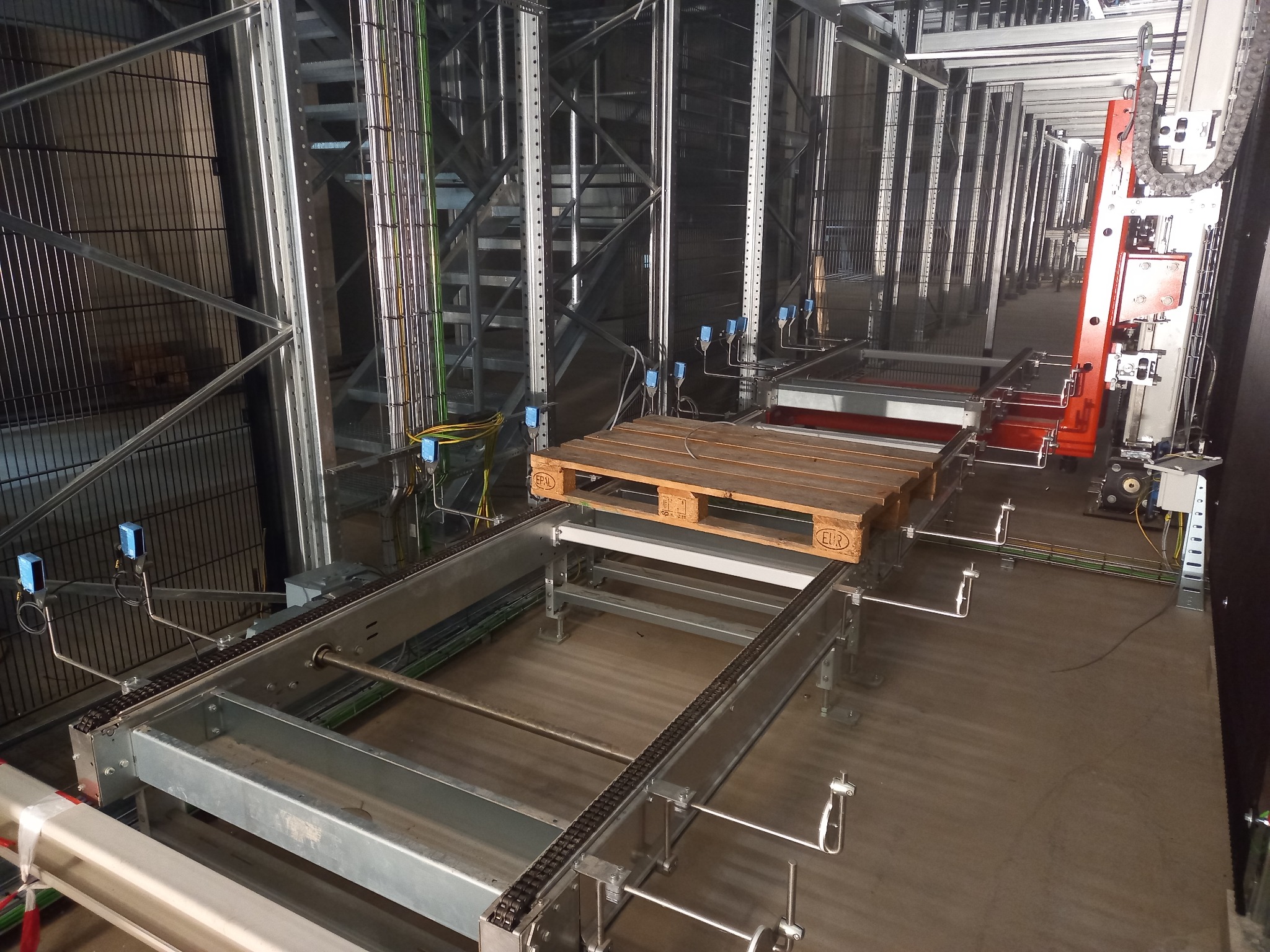 Automatyczny system transportu palet w magazynie wysokiego składowania. Widoczne metalowe konstrukcje, czujniki i okablowanie. Paleta na platformie transportowej.