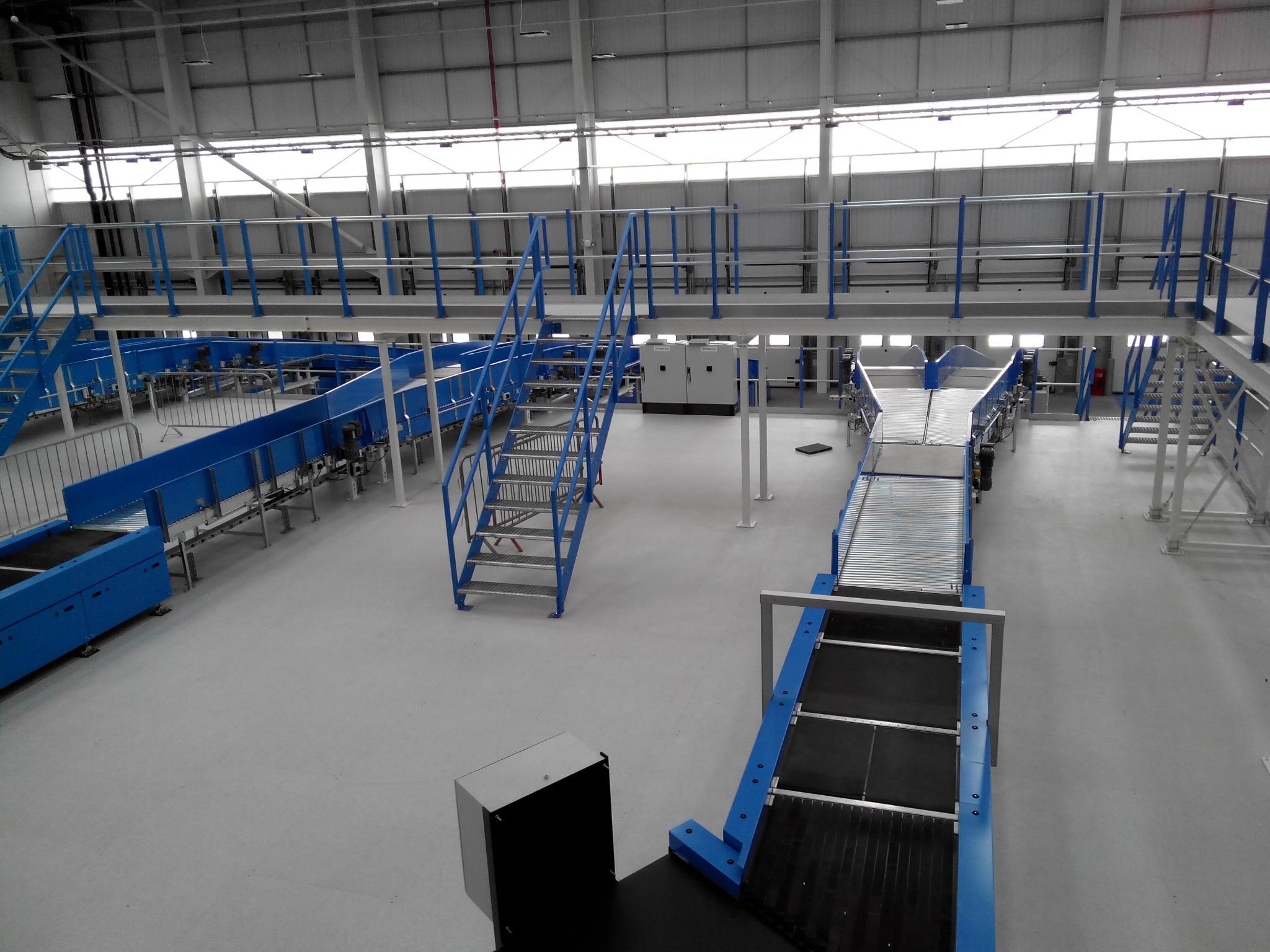 Wnętrze hali produkcyjnej z systemem transportu taśmowego w kolorze niebieskim, widok z góry. Metalowe schody i podesty, biała podłoga i sufit.