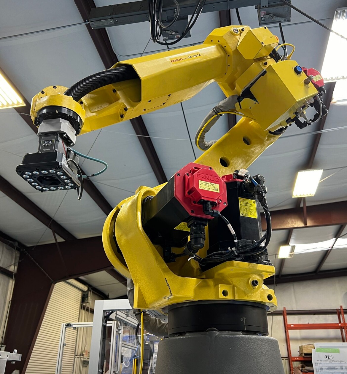 Żółty robot przemysłowy Fanuc M-20iA z czarną głowicą i czerwonymi elementami, widoczny w hali produkcyjnej z metalową konstrukcją dachu.