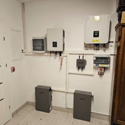 CLEAN&GREEN ADRIAN TOMASZEWSKI - Kompletna instalacja: dwa szare magazyny energii Fox, inwertery na ścianie, skrzynka elektryczna i zabezpieczenia. Widok na jasne pomieszczenie z panelami.
