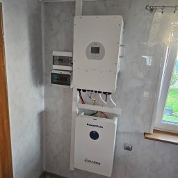 CLEAN&GREEN ADRIAN TOMASZEWSKI - Wnętrze z zamontowanym białym magazynem energii PowerGem i inwerterem Deye na ścianie o marmurkowej fakturze, obok okna z firanką i brązowym parapetem.