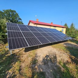 CLEAN&GREEN ADRIAN TOMASZEWSKI - Panel słoneczny na metalowej konstrukcji, na tle domu z czerwoną dachówką i zieleni. Błękitne niebo. Instalacja w wiejskim otoczeniu.