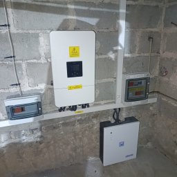 CLEAN&GREEN ADRIAN TOMASZEWSKI - Zamontowany inwerter fotowoltaiczny i magazyn energii na szarej ścianie z betonu komórkowego. Widoczne kable i skrzynki elektryczne. Instalacja w piwnicy.