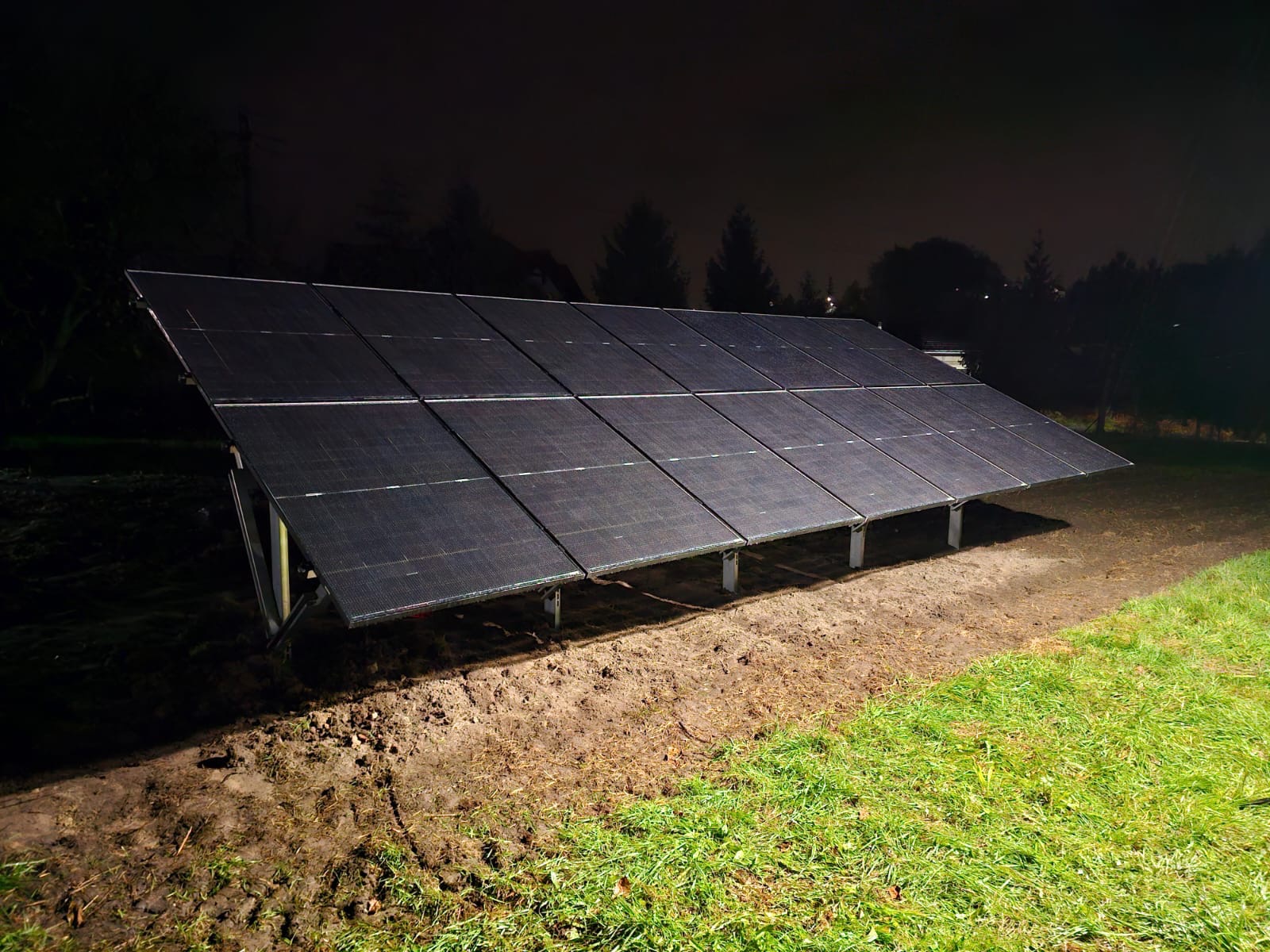 Nocny widok na instalację paneli słonecznych na trawiastym terenie. Oświetlenie podkreśla strukturę paneli i ich metalową konstrukcję. Ciemne tło drzew.