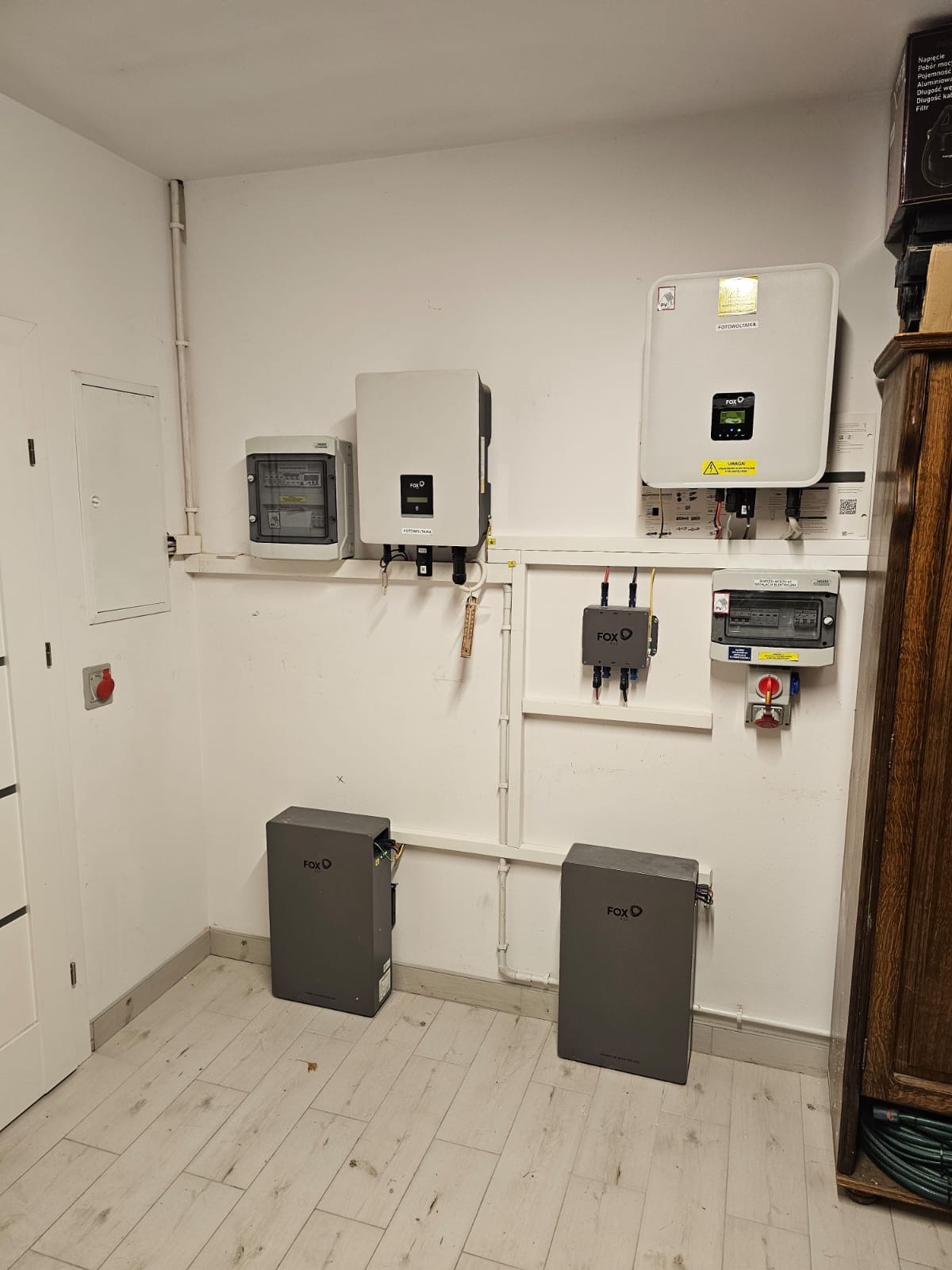 Kompletna instalacja: dwa szare magazyny energii Fox, inwertery na ścianie, skrzynka elektryczna i zabezpieczenia. Widok na jasne pomieszczenie z panelami.