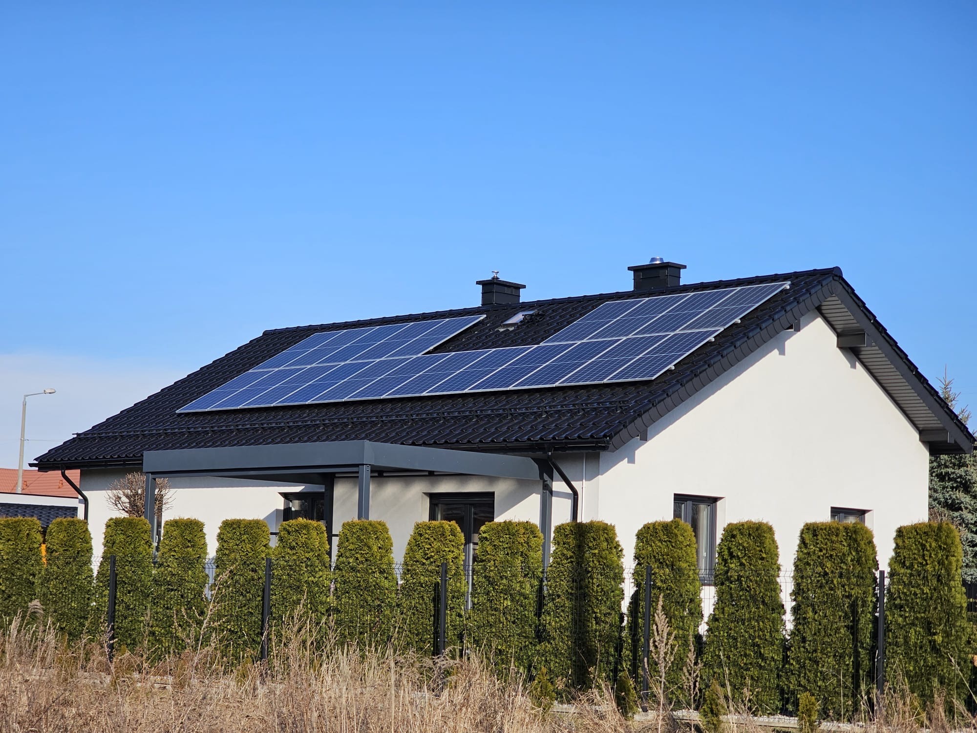 Biały dom jednorodzinny z ciemną dachówką i panelami słonecznymi na dachu, widok zza żywopłotu z tui, błękitne niebo w tle.