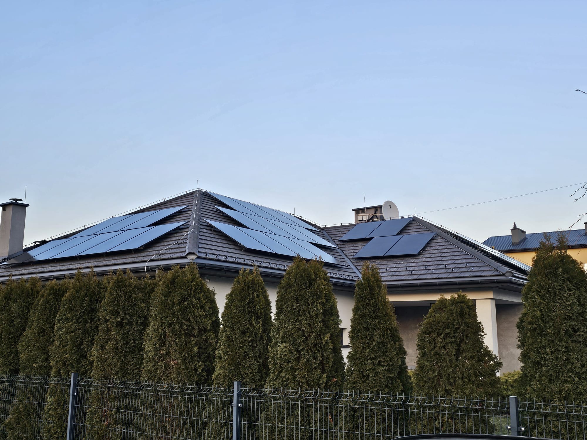 Dom jednorodzinny z panelami słonecznymi na dachu, widoczny zza żywopłotu i metalowego ogrodzenia. Ujęcie z dołu, w tle błękitne niebo.