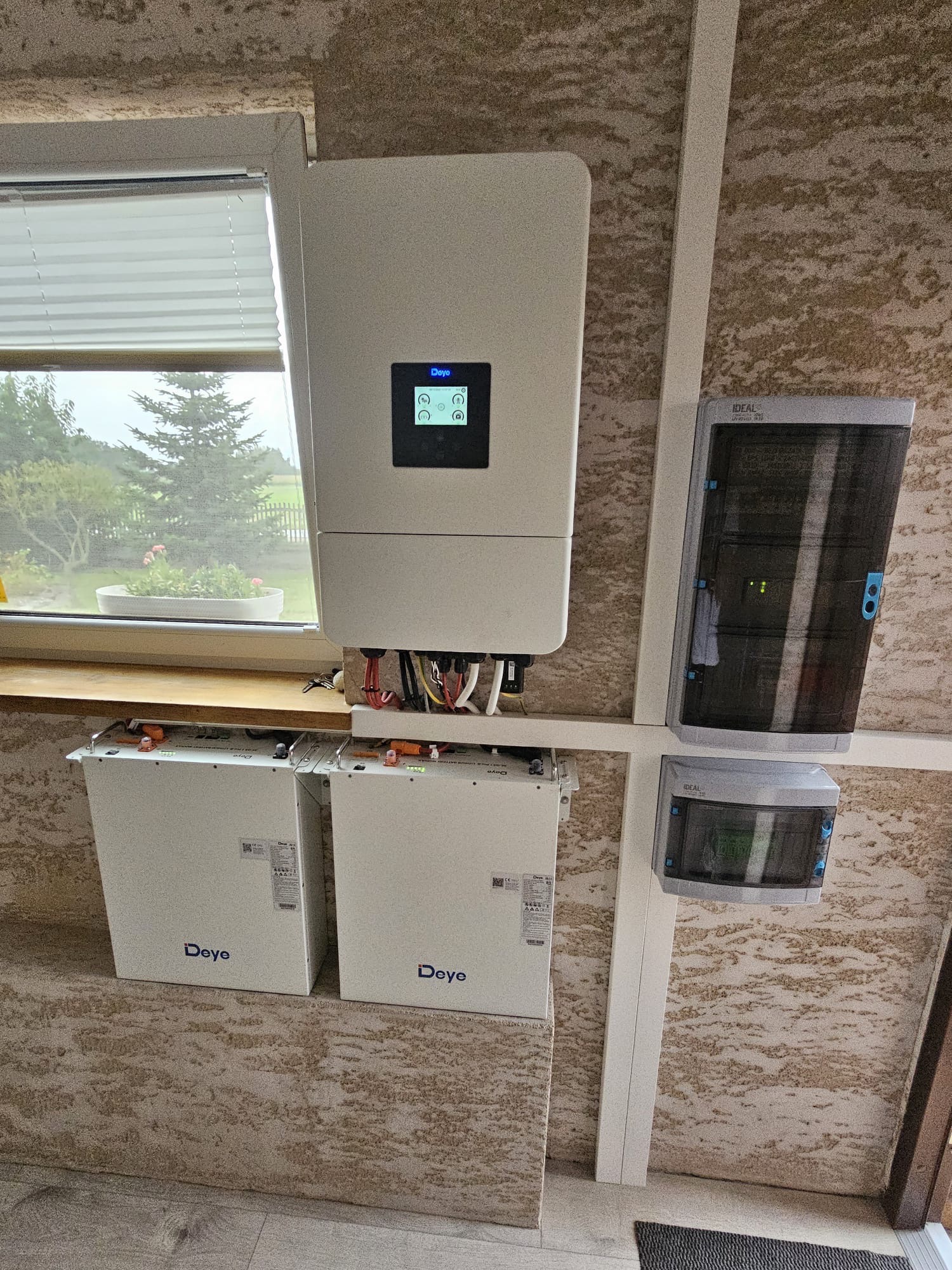 Zamontowany system zasilania awaryjnego Deye z dwoma akumulatorami, inwerterem i rozdzielnicą na ścianie o fakturze baranka, widok z dołu.