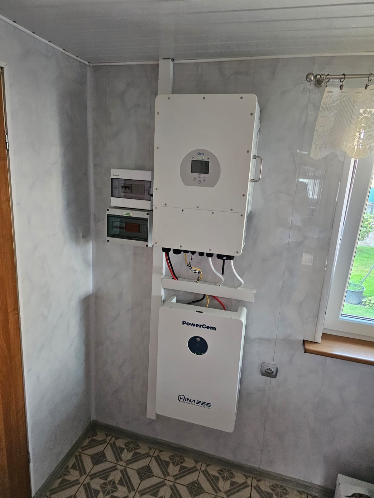 Wnętrze z zamontowanym białym magazynem energii PowerGem i inwerterem Deye na ścianie o marmurkowej fakturze, obok okna z firanką i brązowym parapetem.