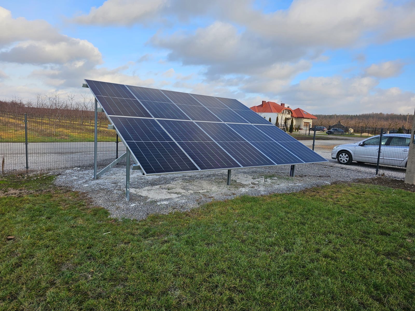 Instalacja paneli słonecznych na trawniku, widok z dołu, w tle dom z czerwoną dachówką i srebrny samochód za ogrodzeniem. Błękitne niebo z chmurami.