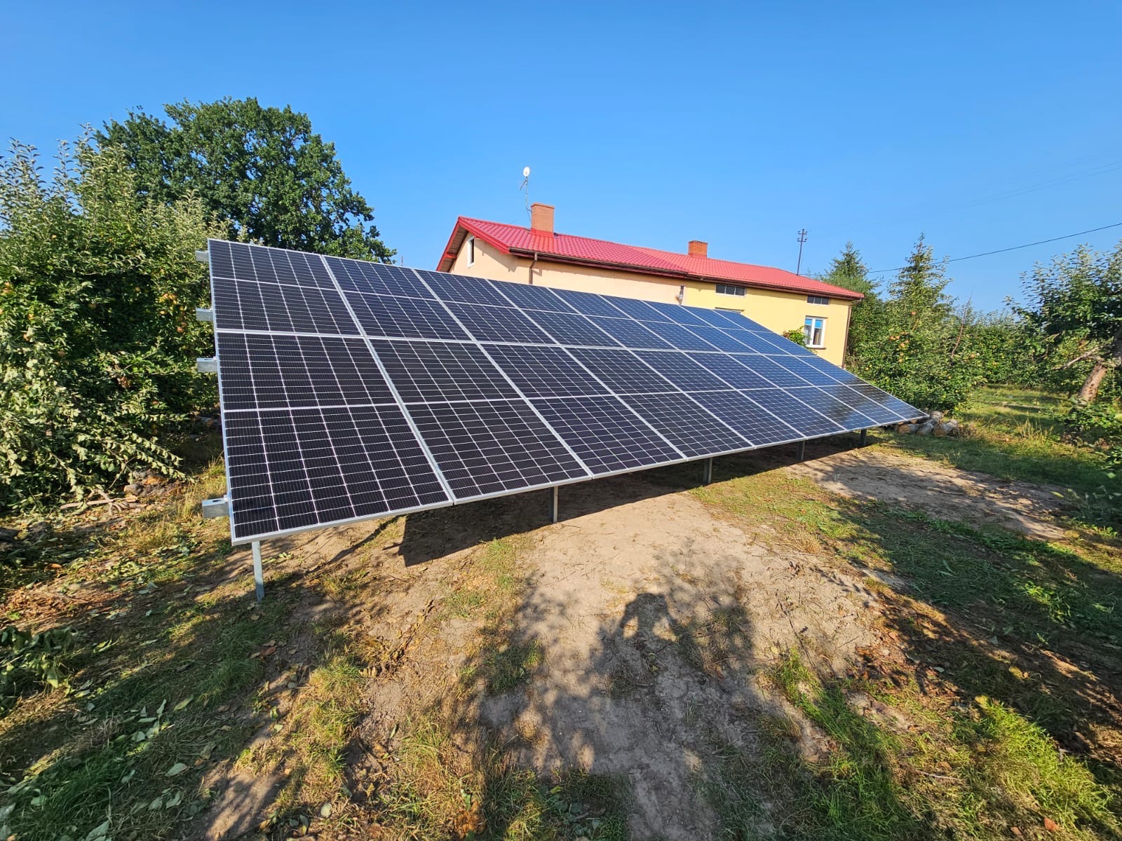 Panel słoneczny na metalowej konstrukcji, na tle domu z czerwoną dachówką i zieleni. Błękitne niebo. Instalacja w wiejskim otoczeniu.