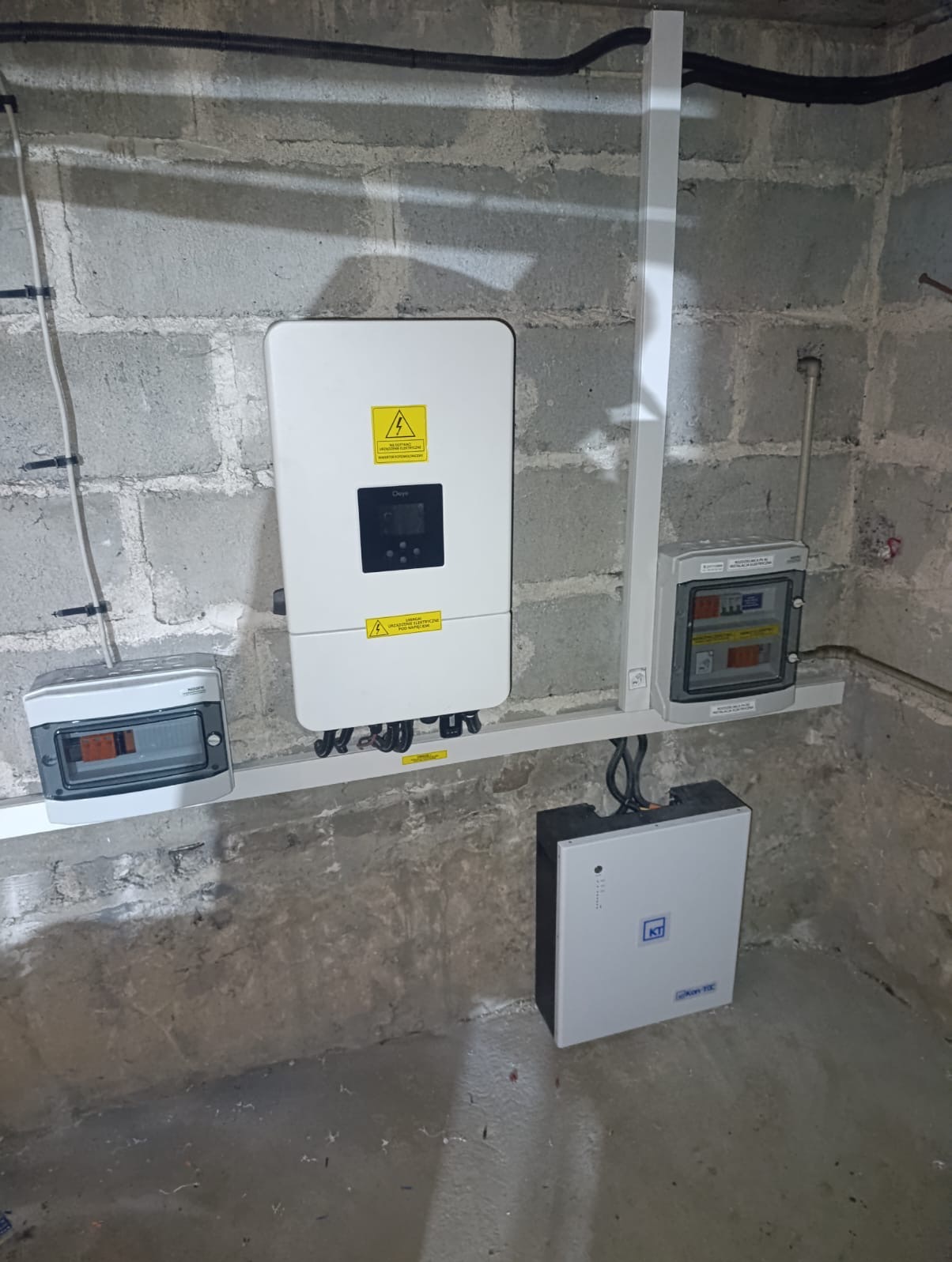 Zamontowany inwerter fotowoltaiczny i magazyn energii na szarej ścianie z betonu komórkowego. Widoczne kable i skrzynki elektryczne. Instalacja w piwnicy.