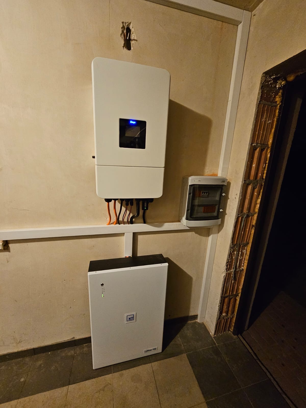 Biały inwerter z podłączonymi kablami i skrzynką elektryczną na ścianie, obok akumulator energii na podłodze. Widoczne elementy instalacji wewnątrz budynku.