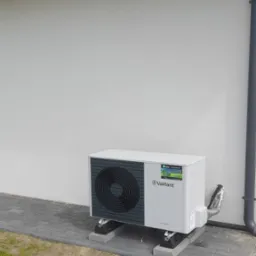 Biała pompa ciepła Vaillant zamontowana na betonowych bloczkach przy ścianie budynku z drewnianą podbitką i rynną spustową.