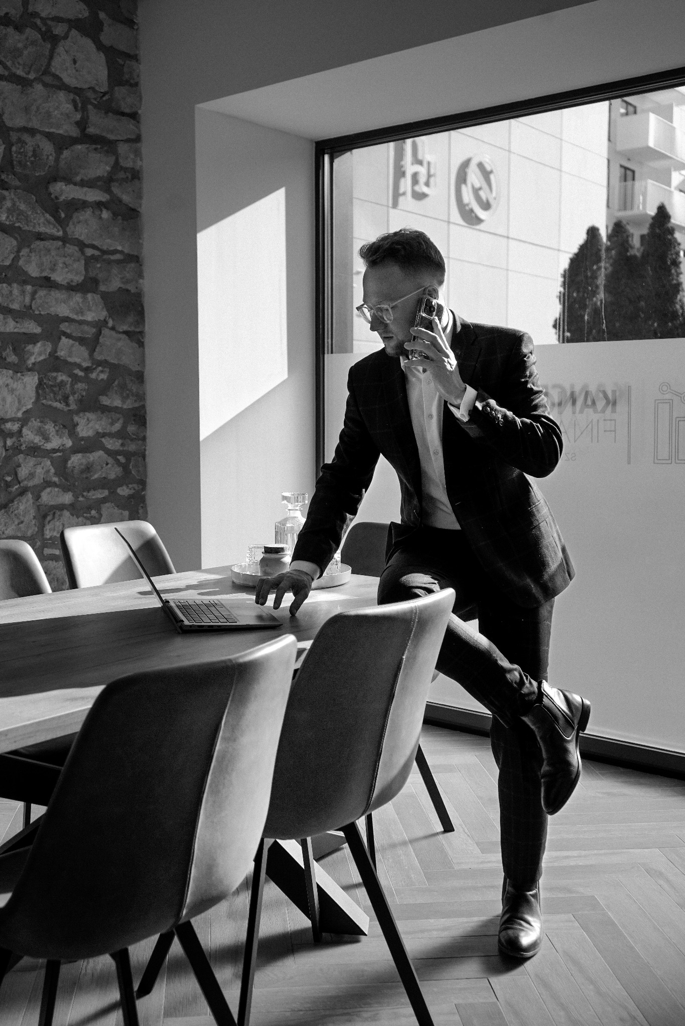 Czarno-białe zdjęcie mężczyzny w garniturze, rozmawiającego przez telefon i pracującego na laptopie przy drewnianym stole w nowoczesnym biurze z kamienną ścianą i dużym oknem.