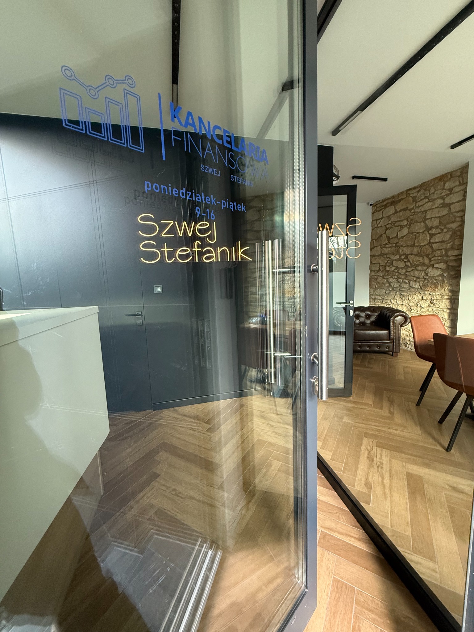 Nowoczesne biuro Kancelarii Finansowej Szwej Stefanik z przeszklonym wejściem, widoczne logo i godziny otwarcia. Wnętrze z kamienną ścianą i skórzanym fotelem.