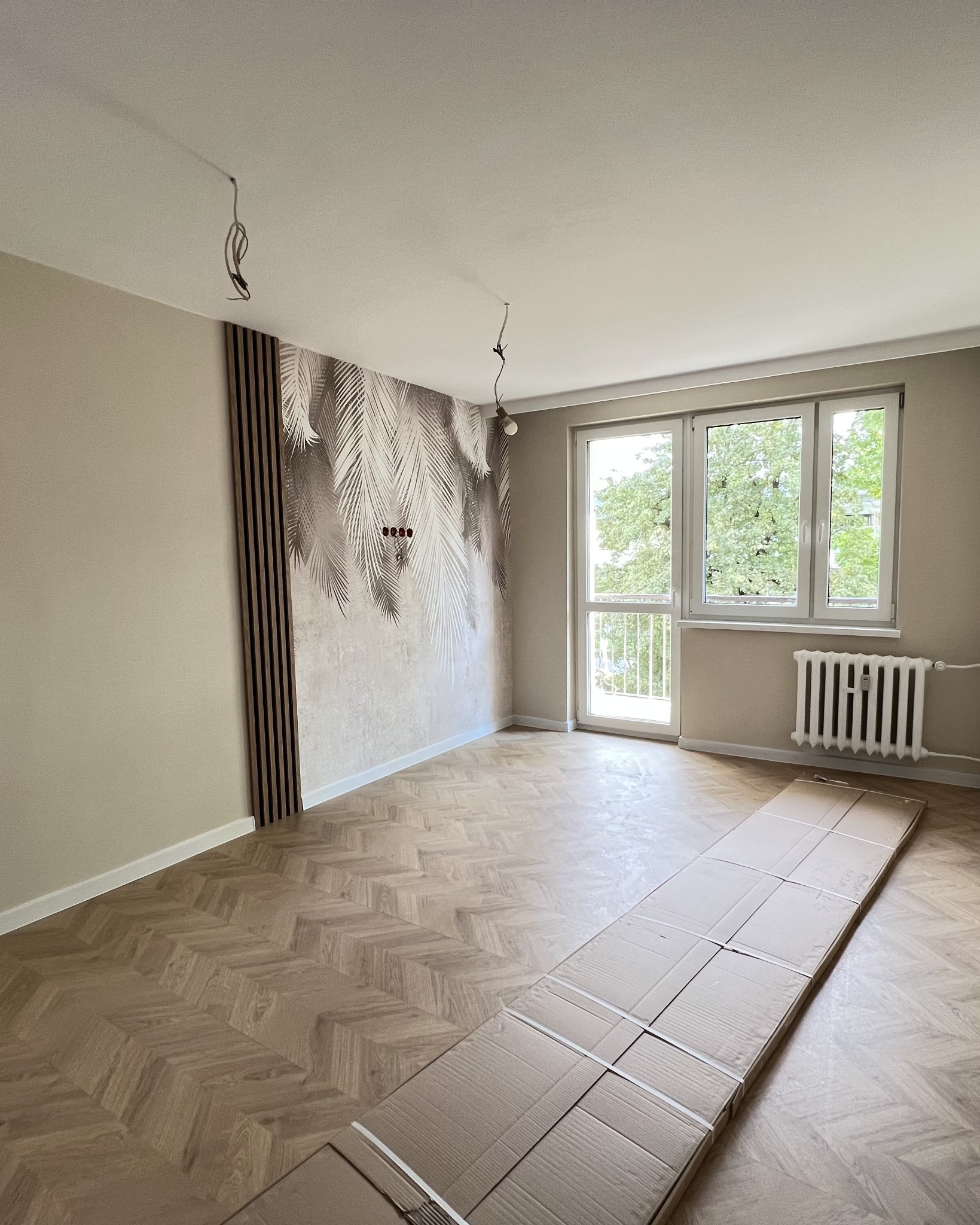 Pokój po remoncie: parkiet w jodełkę, ściana z motywem liści palmowych i drewniane lamele. Widoczne kable od lampy i kartony na podłodze.