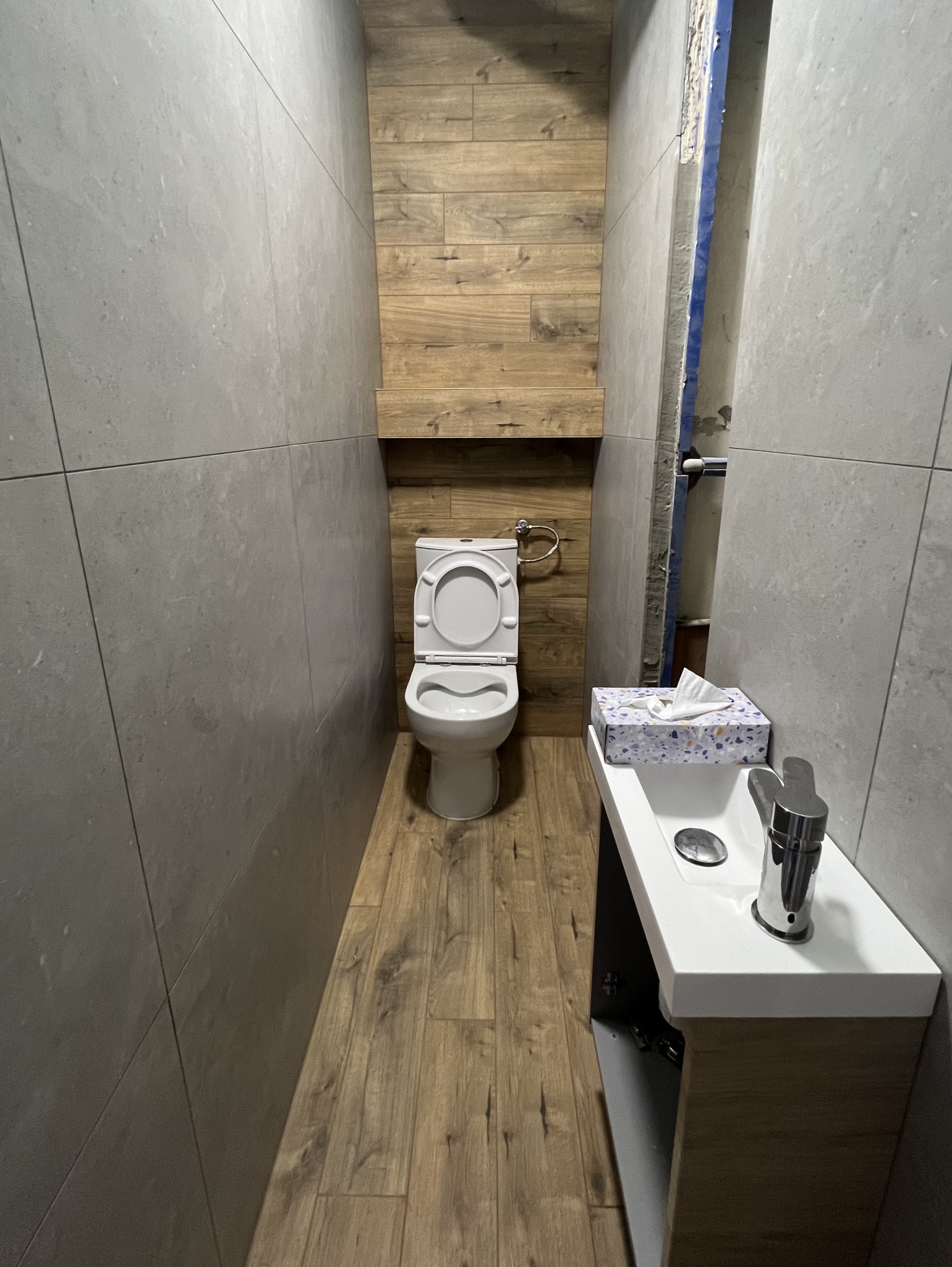 Wąska łazienka po remoncie: szare płytki ścienne, drewnopodobna podłoga i ściana za toaletą, biała umywalka z szafką. Widoczne rury w rogu.