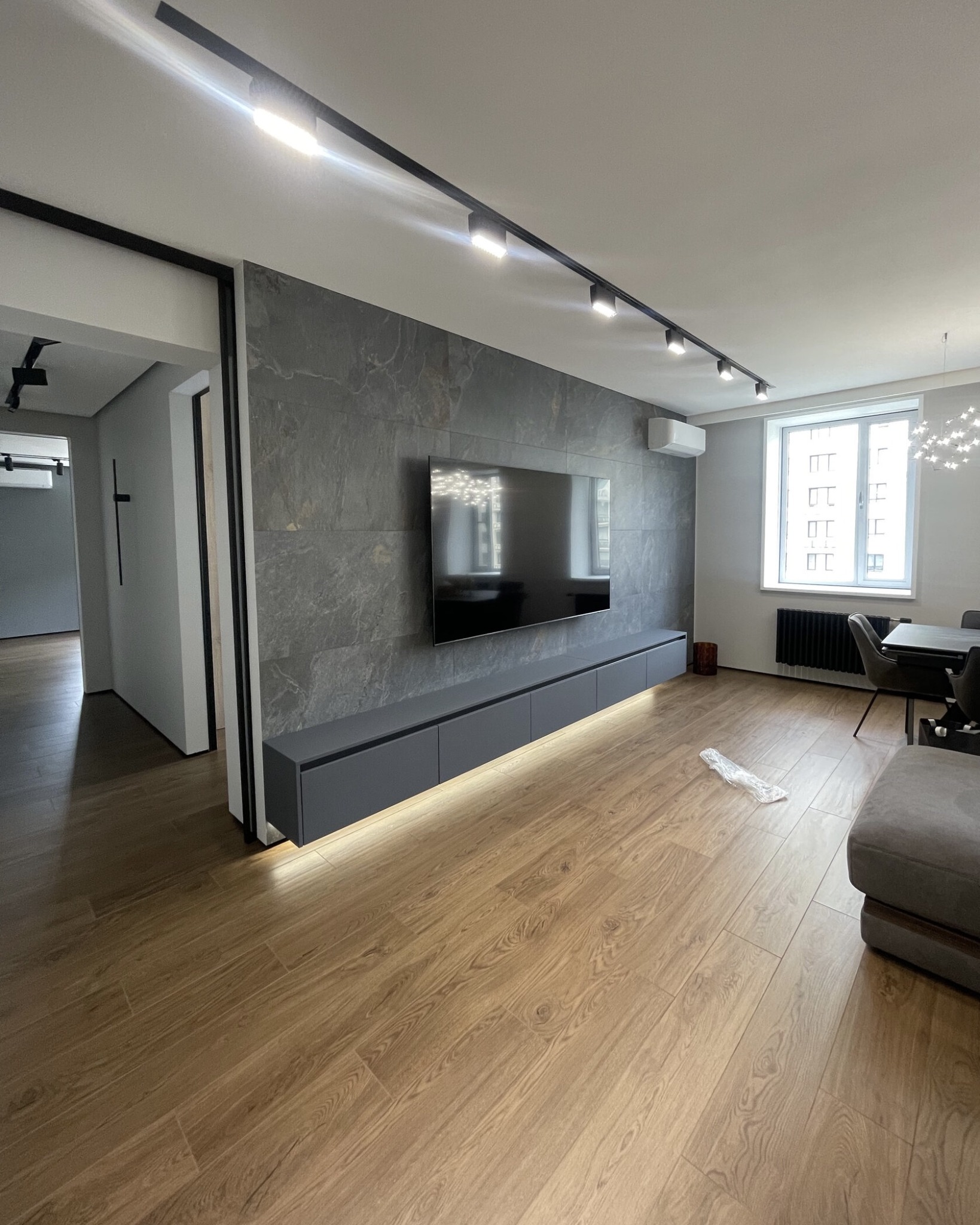 Nowoczesne wnętrze: szara ściana z TV, podświetlana szafka RTV, drewniana podłoga, okno z grzejnikiem i designerskie oświetlenie sufitowe.